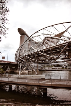 A futuristic bridge with a complex metallic structure spans over a body of water. In the background, a modern architectural building with a unique ship-like design at its top towers above the other structures.