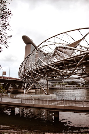 A futuristic bridge with a complex metallic structure spans over a body of water. In the background, a modern architectural building with a unique ship-like design at its top towers above the other structures.
