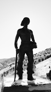 A statue of a rugged miner stands on a snow-covered pedestal, holding a pickaxe and wearing traditional mining attire, including a hat. The background includes a rocky, mountainous landscape under a clear sky.