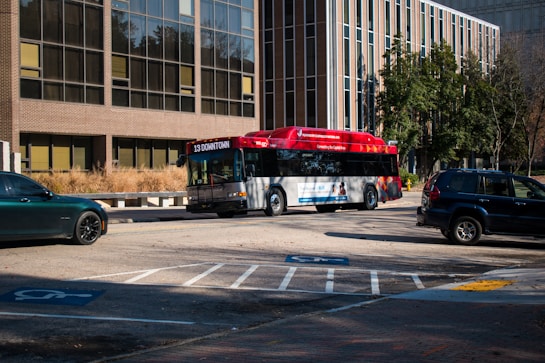 An urban scene featuring a red and grey public transport bus marked with '13 Downtown', driving on a street with parking spaces that include designated handicap spots. The background consists of a large office building with numerous windows and a small number of trees, suggesting a city setting. Several cars are parked nearby, including a dark green and a dark blue vehicle.