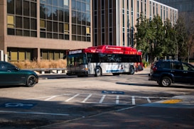 An urban scene featuring a red and grey public transport bus marked with '13 Downtown', driving on a street with parking spaces that include designated handicap spots. The background consists of a large office building with numerous windows and a small number of trees, suggesting a city setting. Several cars are parked nearby, including a dark green and a dark blue vehicle.