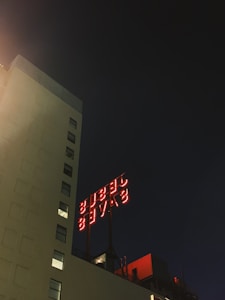 A tall, light-colored building is illuminated against a dark night sky. A large red neon sign on the rooftop reads 'Jesus Saves,' casting a warm glow.