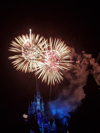 Bright, colorful fireworks explode in the night sky above a beautifully lit castle, which is illuminated in blue and purple hues.