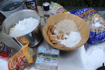 A variety of baking ingredients are arranged on a metallic surface. A large bowl contains oats, cubes of caramel, and white powder, likely flour or powdered sugar. Next to it is a large metal pot with similar white powder. Surrounding the containers are packs of oats, organic salted butter, caramel candies, and a large bag of nuts. A canister of Quaker oats and a salt grinder are also visible.