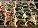 A collection of assorted wraps are neatly arranged in a cardboard box. Each wrap is filled with a variety of fresh ingredients, including leafy greens, sliced meats, and colorful vegetables. The wraps are sliced to reveal cross-sections of their fillings, showcasing layers of lettuce, spinach, slices of red and yellow bell peppers, and thinly sliced onions.