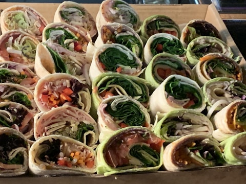 A collection of assorted wraps are neatly arranged in a cardboard box. Each wrap is filled with a variety of fresh ingredients, including leafy greens, sliced meats, and colorful vegetables. The wraps are sliced to reveal cross-sections of their fillings, showcasing layers of lettuce, spinach, slices of red and yellow bell peppers, and thinly sliced onions.