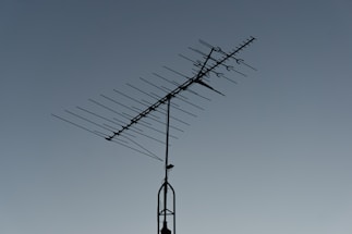 A rooftop television antenna stands silhouetted against a clear, twilight sky. The antenna's multiple metal rods are aligned in a structured pattern, and a small bird is perched on the upper section.
