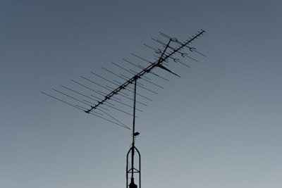 A rooftop television antenna stands silhouetted against a clear, twilight sky. The antenna's multiple metal rods are aligned in a structured pattern, and a small bird is perched on the upper section.