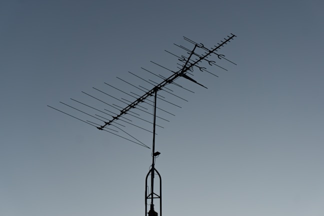 A rooftop television antenna stands silhouetted against a clear, twilight sky. The antenna's multiple metal rods are aligned in a structured pattern, and a small bird is perched on the upper section.