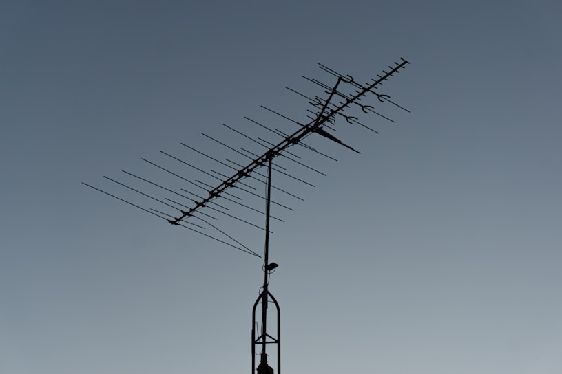 A rooftop television antenna stands silhouetted against a clear, twilight sky. The antenna's multiple metal rods are aligned in a structured pattern, and a small bird is perched on the upper section.