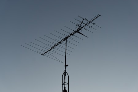 A rooftop television antenna stands silhouetted against a clear, twilight sky. The antenna's multiple metal rods are aligned in a structured pattern, and a small bird is perched on the upper section.