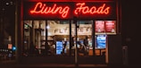 A brightly lit restaurant interior is visible through large windows. The neon sign 'Living Foods' in red lettering stands out prominently. Inside, several people are seated and involved in conversations or activities. Shelves and coolers with food and drinks can be seen, along with a menu board displaying options like salads and quinoa bowls.