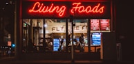 A brightly lit restaurant interior is visible through large windows. The neon sign 'Living Foods' in red lettering stands out prominently. Inside, several people are seated and involved in conversations or activities. Shelves and coolers with food and drinks can be seen, along with a menu board displaying options like salads and quinoa bowls.