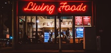 A brightly lit restaurant interior is visible through large windows. The neon sign 'Living Foods' in red lettering stands out prominently. Inside, several people are seated and involved in conversations or activities. Shelves and coolers with food and drinks can be seen, along with a menu board displaying options like salads and quinoa bowls.