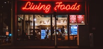 A brightly lit restaurant interior is visible through large windows. The neon sign 'Living Foods' in red lettering stands out prominently. Inside, several people are seated and involved in conversations or activities. Shelves and coolers with food and drinks can be seen, along with a menu board displaying options like salads and quinoa bowls.