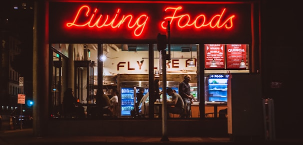 A brightly lit restaurant interior is visible through large windows. The neon sign 'Living Foods' in red lettering stands out prominently. Inside, several people are seated and involved in conversations or activities. Shelves and coolers with food and drinks can be seen, along with a menu board displaying options like salads and quinoa bowls.