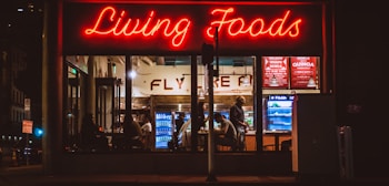A brightly lit restaurant interior is visible through large windows. The neon sign 'Living Foods' in red lettering stands out prominently. Inside, several people are seated and involved in conversations or activities. Shelves and coolers with food and drinks can be seen, along with a menu board displaying options like salads and quinoa bowls.
