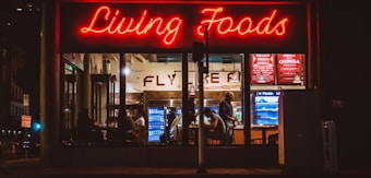 A brightly lit restaurant interior is visible through large windows. The neon sign 'Living Foods' in red lettering stands out prominently. Inside, several people are seated and involved in conversations or activities. Shelves and coolers with food and drinks can be seen, along with a menu board displaying options like salads and quinoa bowls.