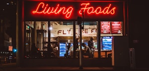 A brightly lit restaurant interior is visible through large windows. The neon sign 'Living Foods' in red lettering stands out prominently. Inside, several people are seated and involved in conversations or activities. Shelves and coolers with food and drinks can be seen, along with a menu board displaying options like salads and quinoa bowls.