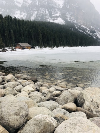 A serene natural landscape featuring a partially frozen lake surrounded by pine forests and snow-capped mountains. In the foreground, large smooth rocks line the lakeshore. A rustic wooden cabin is nestled among the trees on the left side.