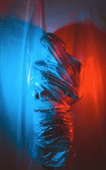 A figure is wrapped in a translucent plastic material, with contrasting blue and red lighting casting colorful shadows and reflections. The printed text on the plastic creates an abstract and slightly industrial appearance.