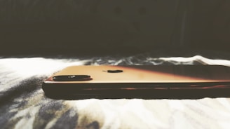 A smartphone with a sleek, metallic finish is lying on a soft, textured surface, illuminated by natural light that casts soft shadows.