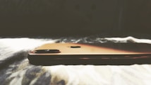 A smartphone with a sleek, metallic finish is lying on a soft, textured surface, illuminated by natural light that casts soft shadows.