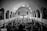 A large congregation gathers inside a grand church with a high arched ceiling and multiple arches along the sides. The focus is towards a central cross at the altar, with people standing and sitting, creating a lively and engaging atmosphere. The image is in black and white, emphasizing contrasts and architectural details.