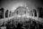 A large congregation gathers inside a grand church with a high arched ceiling and multiple arches along the sides. The focus is towards a central cross at the altar, with people standing and sitting, creating a lively and engaging atmosphere. The image is in black and white, emphasizing contrasts and architectural details.