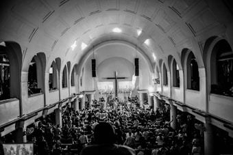 A large congregation gathers inside a grand church with a high arched ceiling and multiple arches along the sides. The focus is towards a central cross at the altar, with people standing and sitting, creating a lively and engaging atmosphere. The image is in black and white, emphasizing contrasts and architectural details.