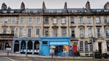 A row of historic stone buildings with multiple businesses at street level. Notable features include a blue-colored bookstore with a bright facade and several bay windows in the upper stories. The architecture is characterized by classical design with uniform windows and chimneys.