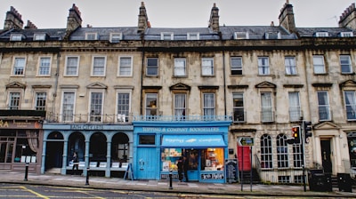 A row of historic stone buildings with multiple businesses at street level. Notable features include a blue-colored bookstore with a bright facade and several bay windows in the upper stories. The architecture is characterized by classical design with uniform windows and chimneys.