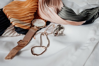 A gold wristwatch with a white face is placed alongside a pair of gold wire glasses, a large gemstone ring, and a pleated scarf featuring shades of orange, pink, black, and green. The items are laid out on a crisp white shirt.