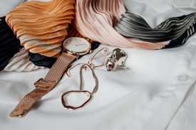 A gold wristwatch with a white face is placed alongside a pair of gold wire glasses, a large gemstone ring, and a pleated scarf featuring shades of orange, pink, black, and green. The items are laid out on a crisp white shirt.