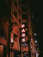 The image features the corner of a building at night with a vertical neon sign displaying the word 'PARK' in red lights. The building's facade has a geometric, textured design, and there is a smaller sign at the bottom that reads 'PMI' and 'New York Convenience'. The overall ambiance is dimly lit with an urban, nighttime setting.
