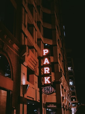 The image features the corner of a building at night with a vertical neon sign displaying the word 'PARK' in red lights. The building's facade has a geometric, textured design, and there is a smaller sign at the bottom that reads 'PMI' and 'New York Convenience'. The overall ambiance is dimly lit with an urban, nighttime setting.