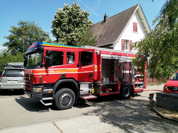 A red fire truck is parked on a residential street next to a white house with brown roof shingles. The truck has open compartments displaying equipment, and is surrounded by trees providing greenery and shade. Nearby, there are other vehicles, including a silver SUV and a red car. The setting appears calm and residential.