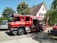 A red fire truck is parked on a residential street next to a white house with brown roof shingles. The truck has open compartments displaying equipment, and is surrounded by trees providing greenery and shade. Nearby, there are other vehicles, including a silver SUV and a red car. The setting appears calm and residential.