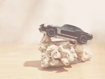 A toy car is precariously balanced on a rough, coral-like rock structure. The setting is minimalistic with a neutral, soft background that emphasizes the vintage appearance of the car.