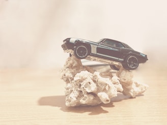 A toy car is precariously balanced on a rough, coral-like rock structure. The setting is minimalistic with a neutral, soft background that emphasizes the vintage appearance of the car.