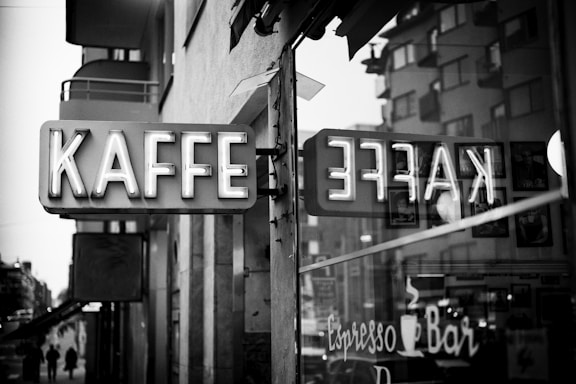 A vintage-style vertical neon sign displaying the word 'KAFFE' is mounted on an exterior wall, with its reflection visible in a window. The black and white image captures an urban setting, with buildings and a couple of silhouetted pedestrians in the background. Inside the window, part of a café interior can be seen with pictures and signage for an espresso bar.