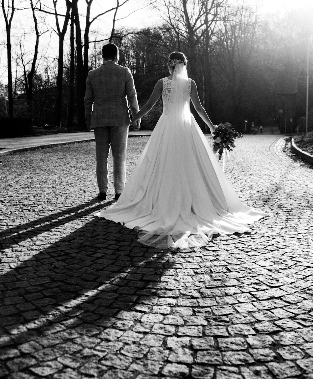 grayscale photo of newly weds walking while holding hands near bare trees