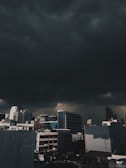 Dark storm clouds loom ominously over a cityscape comprised of various modern buildings. The heavy clouds create a dramatic contrast with the structures below, casting shadows and indicating an impending storm.
