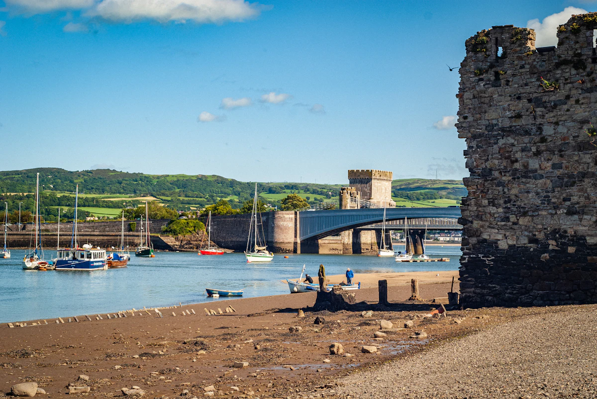 Conwy harbour at low tide, with an old stone tower, moored boats and the bridges across the estuary