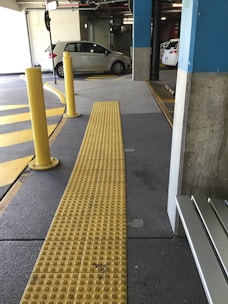 The image features an indoor parking garage with a few parked cars. A yellow tactile paving path for the visually impaired is prominently visible, running through the foreground. Yellow and gray are the dominant colors within the scene. There are also several yellow bollards near the path, and the floor has markings for direction and parking spaces.