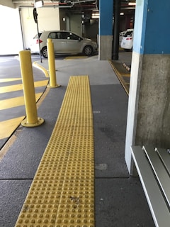 The image features an indoor parking garage with a few parked cars. A yellow tactile paving path for the visually impaired is prominently visible, running through the foreground. Yellow and gray are the dominant colors within the scene. There are also several yellow bollards near the path, and the floor has markings for direction and parking spaces.