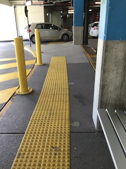 The image features an indoor parking garage with a few parked cars. A yellow tactile paving path for the visually impaired is prominently visible, running through the foreground. Yellow and gray are the dominant colors within the scene. There are also several yellow bollards near the path, and the floor has markings for direction and parking spaces.
