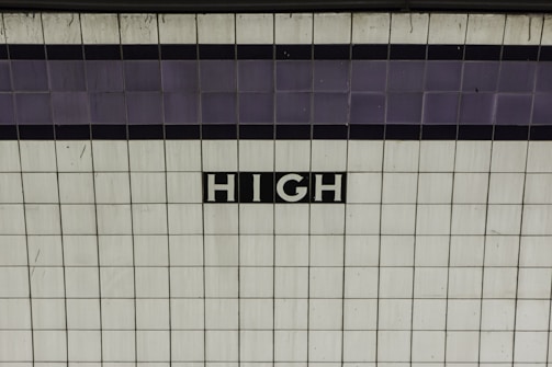 A tiled wall with the word 'HIGH' written in white letters on a black background in the center. The tiles are predominantly white with a subtle pattern of light gray lines. There is a horizontal strip of darker tiles toward the top of the image.