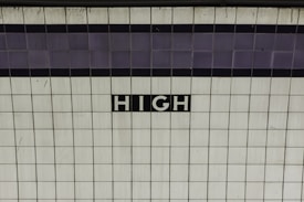 A tiled wall with the word 'HIGH' written in white letters on a black background in the center. The tiles are predominantly white with a subtle pattern of light gray lines. There is a horizontal strip of darker tiles toward the top of the image.