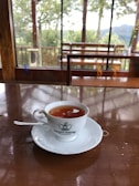A cup of tea is placed on a white saucer atop a polished wooden table. The cup features a logo for Kelani Valley Plantations. Beyond the glass windows, there is a view of green trees and a wooden railing indicating an outdoor landscape.